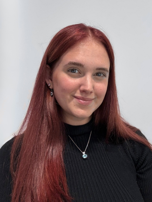 A young woman smiling at the camera with a neutral background behind her. She has red hair and is wearing a black jumper with a pendant on a necklace clearly visible.